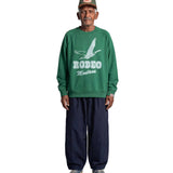 An older man stands against a light gray background wearing a green crewneck sweatshirt with a white 'RODEO Montana' graphic, dark navy pants, and a green cap.