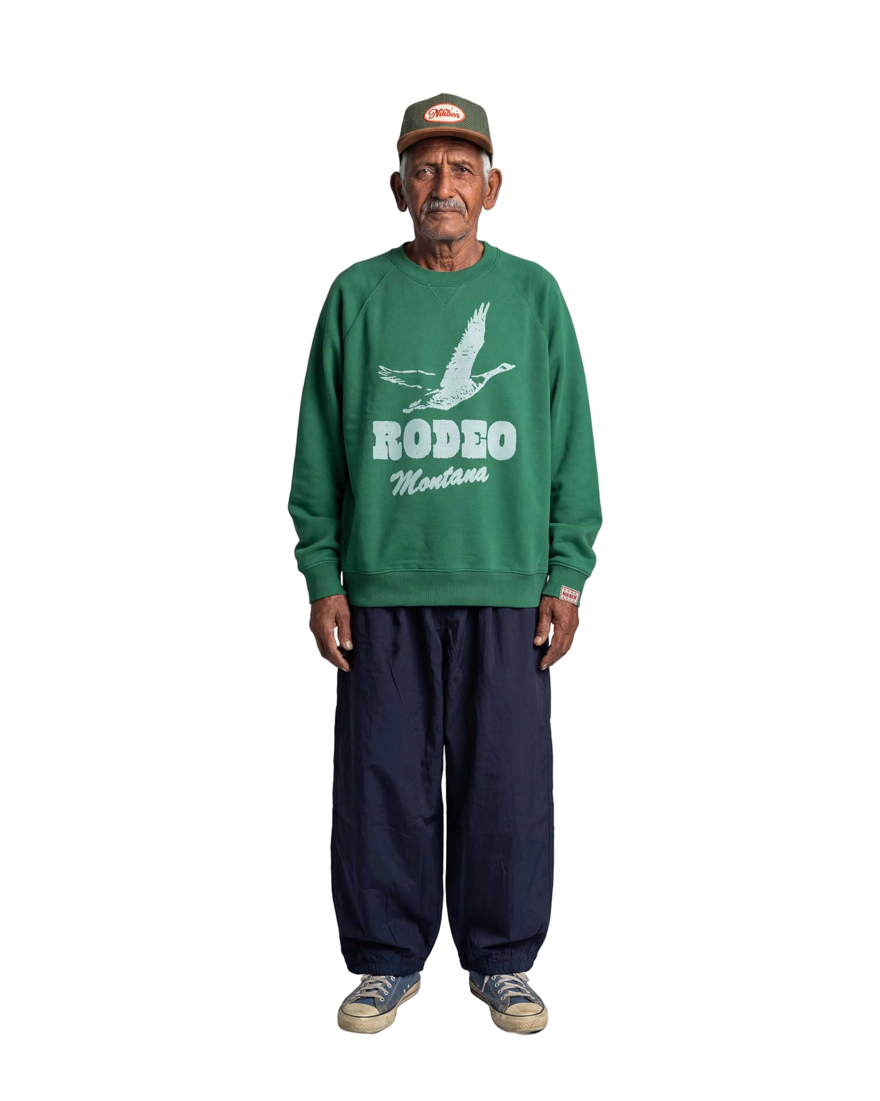 An older man stands against a light gray background wearing a green crewneck sweatshirt with a white 'RODEO Montana' graphic, dark navy pants, and a green cap.