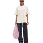 A model stands against a light gray studio background wearing an oversized cream t-shirt with a small black logo on the chest, dark plaid pants, blue sneakers, and holding a fluffy pink bag.