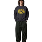 A model stands against a light gray studio background wearing a dark charcoal crewneck sweatshirt with a yellow 'Bottoms Up!' graphic, dark green baggy sweatpants, a pink cap, and pink and blue sneakers.