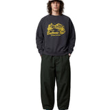 A model stands against a light gray studio background wearing a dark charcoal crewneck sweatshirt with a yellow 'Bottoms Up!' graphic, dark green baggy sweatpants, a pink cap, and pink and blue sneakers.