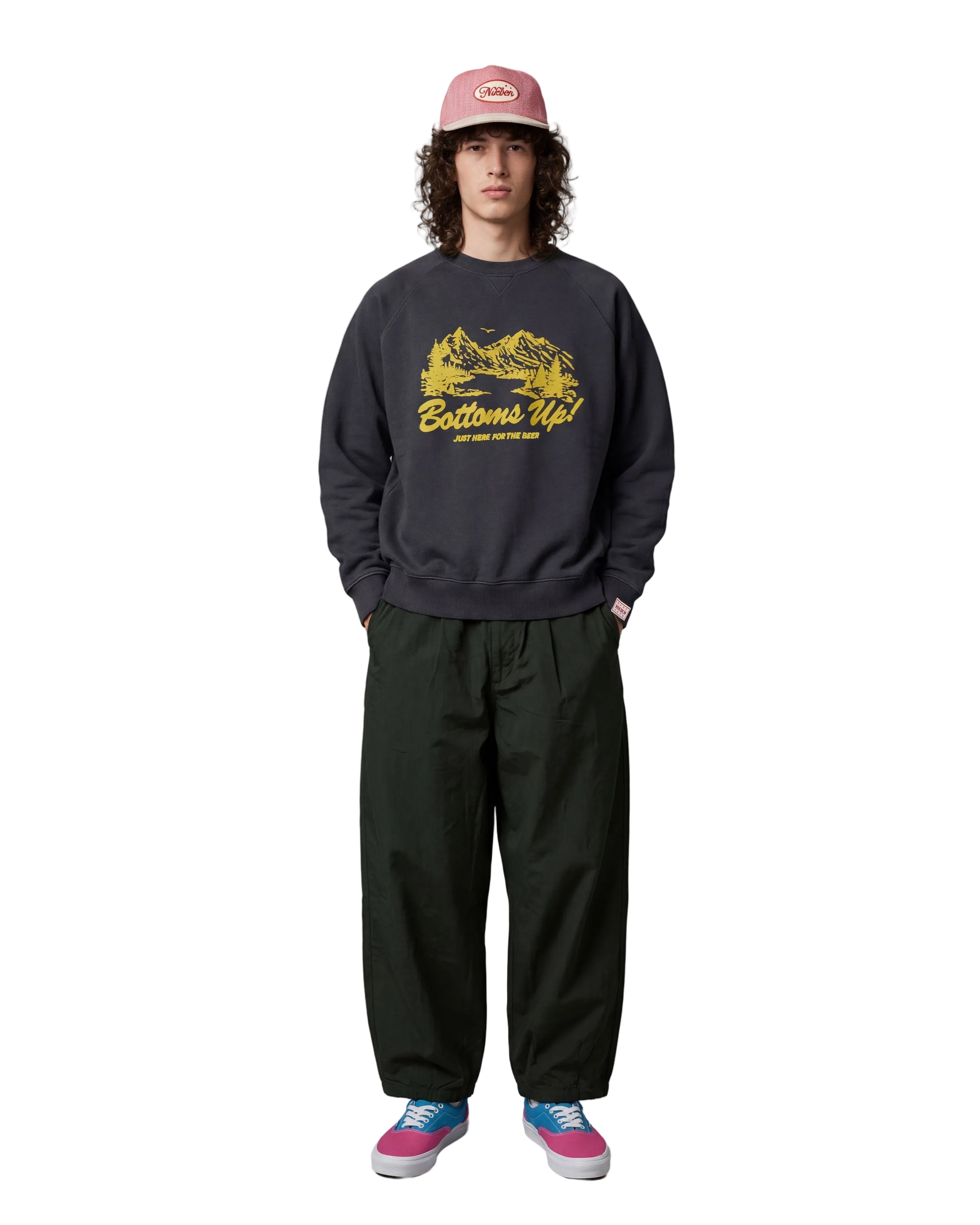 A model stands against a light gray studio background wearing a dark charcoal crewneck sweatshirt with a yellow 'Bottoms Up!' graphic, dark green baggy sweatpants, a pink cap, and pink and blue sneakers.