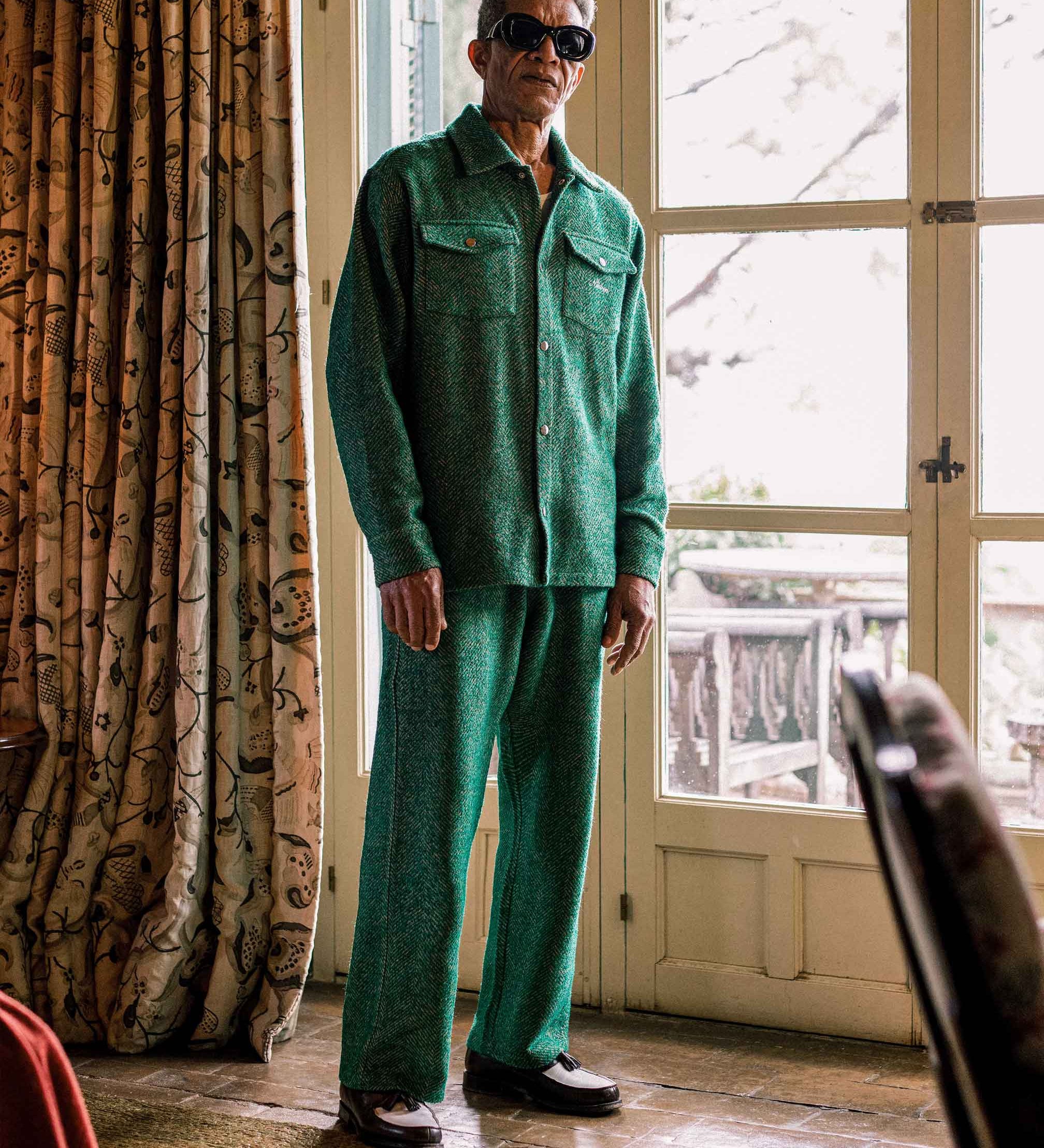 Model is wearing a green outfit standing in a room with a large window and patterned curtain.