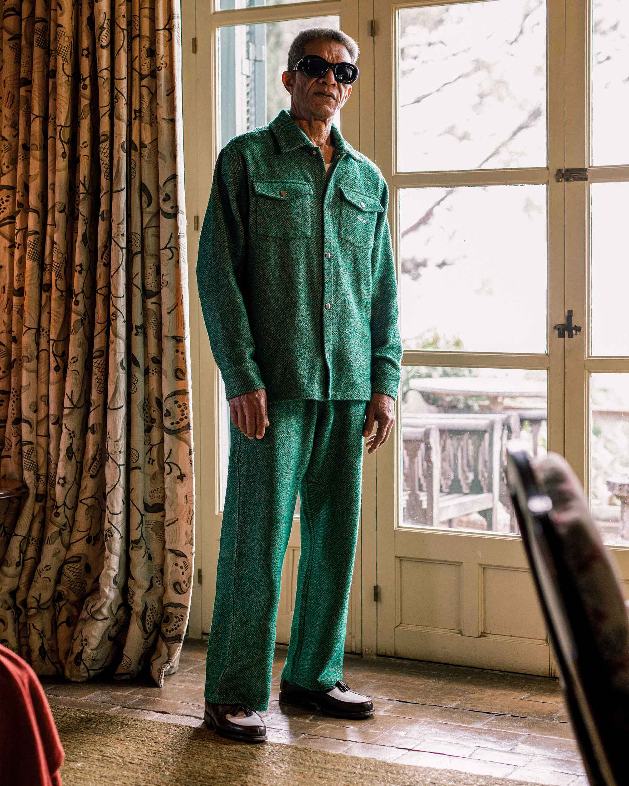Model wearing a green shirt and trouser set standing in a room with curtains and a door.