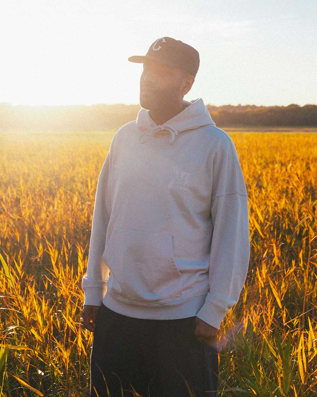 Model wearing a beige hoodie standing in a field with sunset light