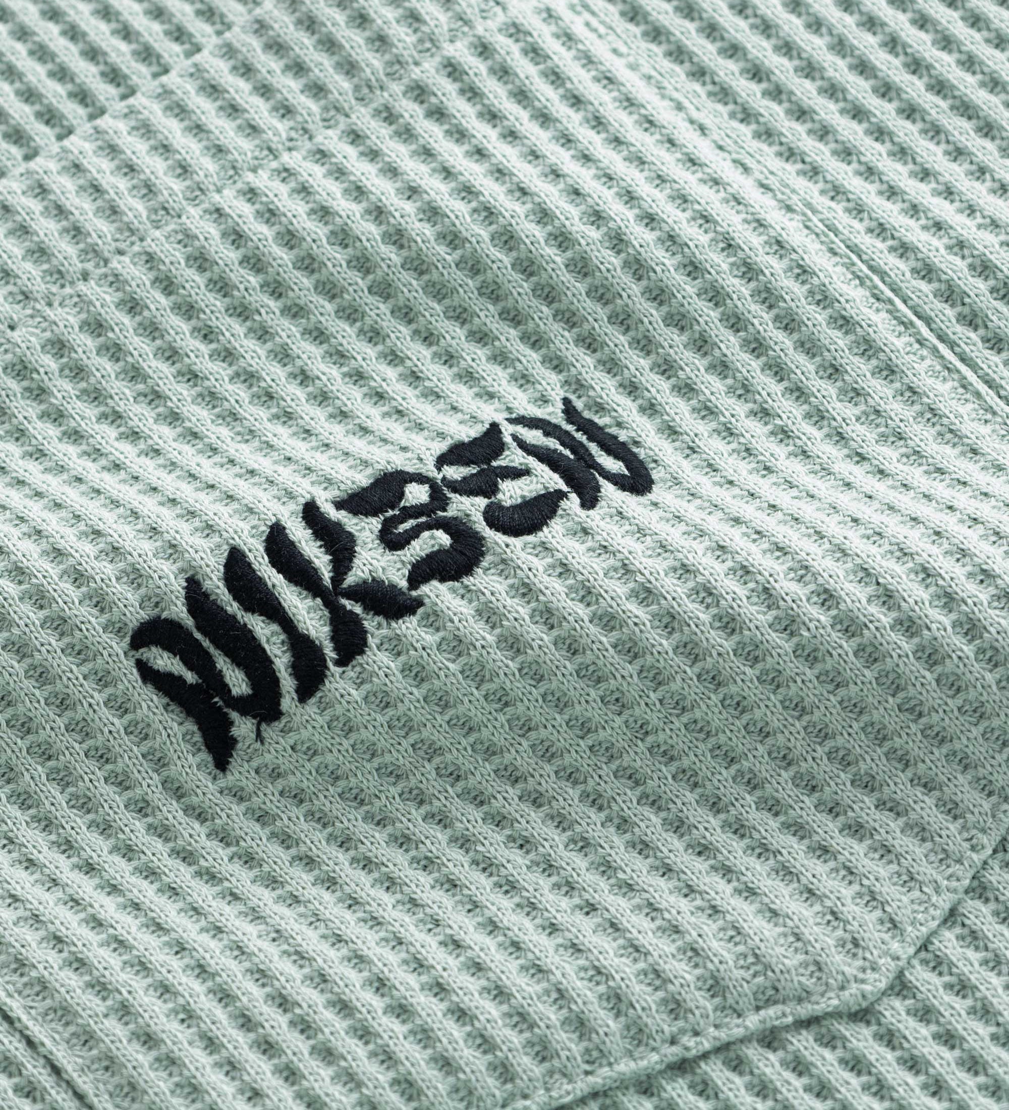 Close-up view of front pocket with stitched black logo on a mint green waffle-patterned shirt